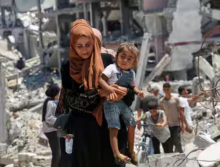 Seorang wanita Palestina menggendong putrinya saat dia berjalan melewati puing-puing rumah yang hancur selama serangan militer Israel, di tengah konflik Israel-Hamas, di Khan Younis di Jalur Gaza selatan 10 Juli 2024 /Reuters